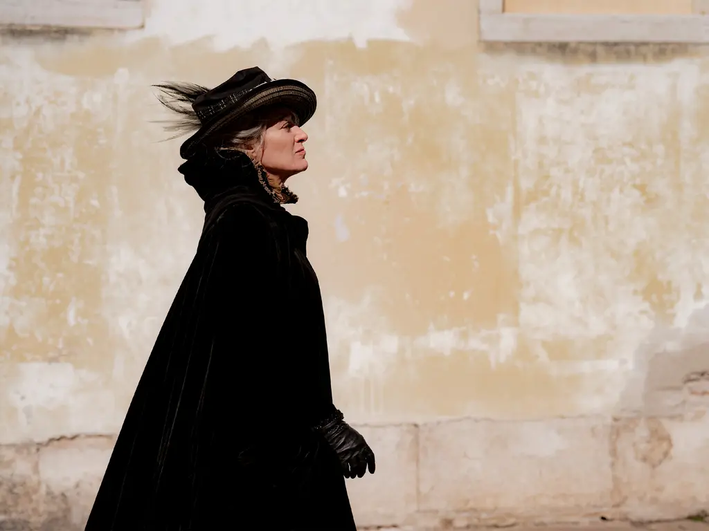 Woman in black coat and hat walking left against pale yellow and white stone wall background.