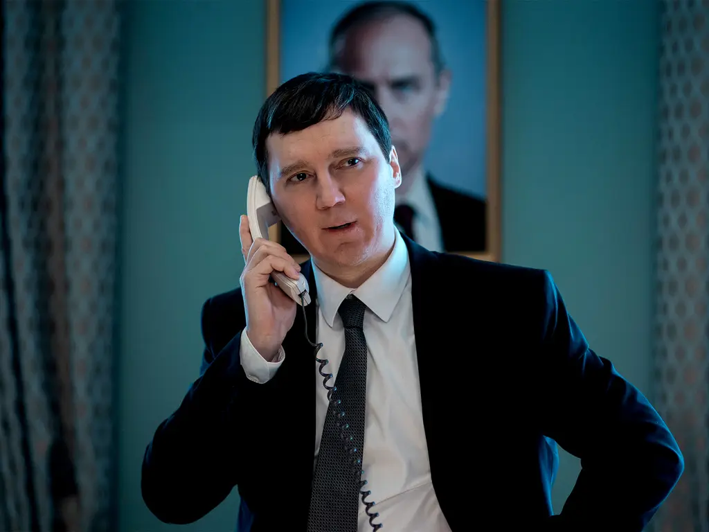 Man in dark suit speaking on phone in formal interior setting, with blurred figure in background.