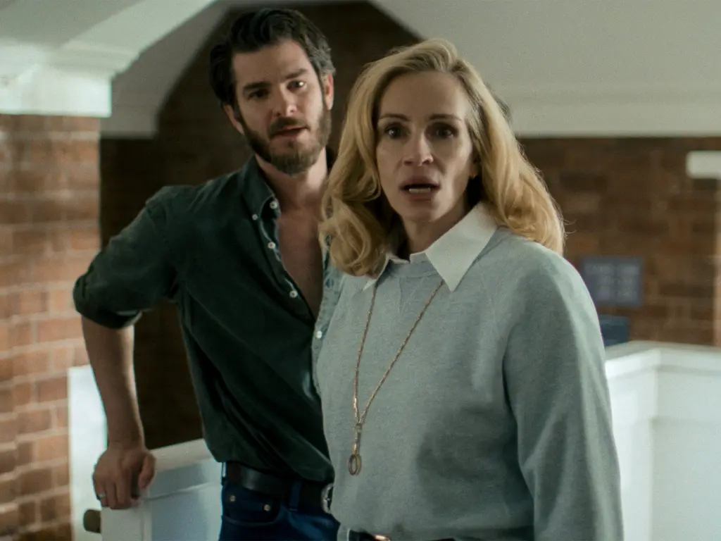 Man in dark green shirt stands behind blonde woman in light blue jumper with white collar in modern kitchen with exposed brick wall.